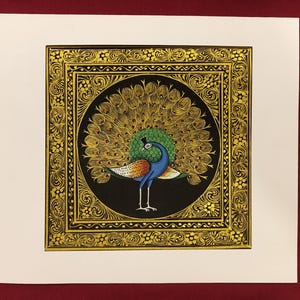 May include: A square painting featuring a central peacock with a vibrant blue body and a fanned tail of gold and green feathers. The peacock is set against a black circular background, enclosed within a detailed gold decorative border on a white mat.