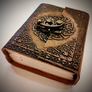 May include: A brown leather journal with a decorative metal clasp and intricate embossed designs. The journal is closed and lying on a white surface.
