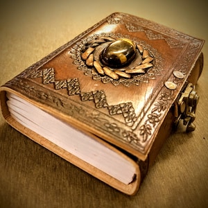May include: A brown leather journal with an intricate embossed design and a decorative clasp. The journal has a gemstone embedded in the front cover.