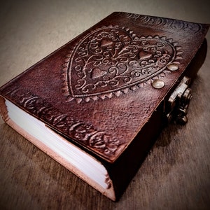 May include: A brown leather journal with a heart-shaped design embossed on the cover. The journal has a metal clasp and is closed.
