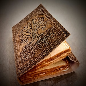 May include: A brown leather-bound journal with a Celtic knot design and a tree of life motif embossed on the cover. The journal is open to reveal blank pages.