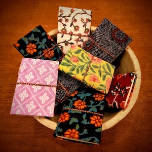 Puede incluir: Una colección de pequeños cuadernos estampados en un cuenco de madera. Los cuadernos presentan varios diseños florales y geométricos en colores como negro, rosa, amarillo y rojo. Cada cuaderno está encuadernado con un cordón de hilo.