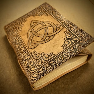 May include: A tan leather journal with a Celtic knot design embossed on the cover. The cover also features ornate corner and border designs. The journal is closed with a string tie and has aged, cream-coloured pages.