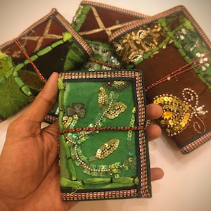 Peut inclure: Carnets faits à la main, richement décorés. Le carnet principal est vert, orné de paillettes dorées et de broderies. D'autres carnets en arrière-plan présentent des accents marron, verts et rouges. Chaque carnet est relié par un cordon décoratif.