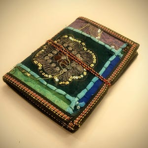 Puede incluir: Un diario de tela multicolor hecho a mano con un diseño floral decorativo. El diario presenta un panel central verde oscuro con lentejuelas doradas, rodeado de parches azules, verdes y morados. Un cordón marrón y rojo envuelve el diario.