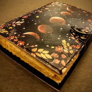 May include: A black leather journal with a floral and moon design. The journal has a clasp closure and aged paper pages.