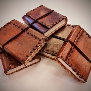 May include: Four brown leather-bound journals with stitching detail and elastic closures. The journals are stacked on top of each other.