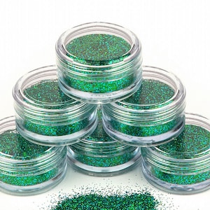 Könnte beinhalten: Sechs kleine, transparente Gläser gefüllt mit grünem Glitter, in Pyramidenform gestapelt. Die Gläser stehen auf einer weißen Oberfläche, und etwas Glitter ist herausgefallen.