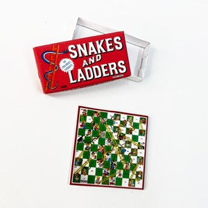 May include: A red box with the words "Snakes and Ladders" on it. The box is open and the game board is visible. The game board is green and white with a checkered pattern. There are snakes and ladders printed on the board.