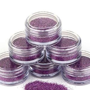 Op de afbeelding: Zes kleine, doorzichtige potjes gevuld met paarse glitter, gestapeld in een piramidevorm. De potjes staan op een wit oppervlak.