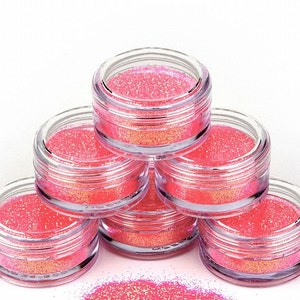 Op de afbeelding: Zes kleine, doorzichtige potjes gevuld met roze glitter, gestapeld in een piramidevorm. De potjes staan op een wit oppervlak.