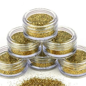 Op de afbeelding: Zes kleine, doorzichtige plastic potjes gevuld met gouden glitter zijn in een piramidevorm gestapeld. Een kleine hoeveelheid glitter is verspreid aan de basis van de potjes. De glitter heeft een fijne textuur en reflecteert licht.