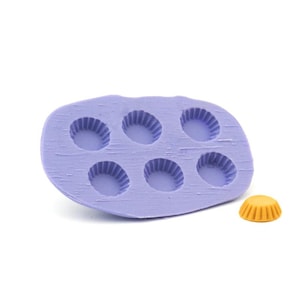 Puede incluir: Molde de silicona morado con seis cavidades redondas, cada una con un borde acanalado. Un pequeño cupcake de silicona amarillo acanalado está al lado del molde.