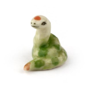 May include: A ceramic snake figurine in shades of green and white, with a red dot on its head. The snake has green spots and a curled body, with a light brown tip on its tail. The figurine is set against a white background.
