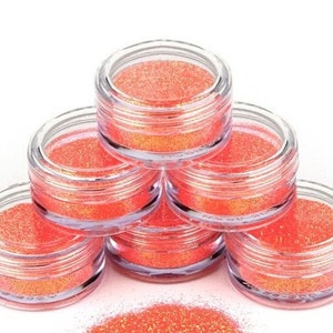 Op de afbeelding: Zes kleine, transparante potjes gevuld met oranje glitter, gestapeld in een piramidevorm. De potjes staan op een wit oppervlak.