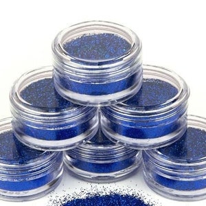 Op de afbeelding: Zes kleine, doorzichtige potjes gevuld met blauwe glitter, gestapeld in een piramidevorm.