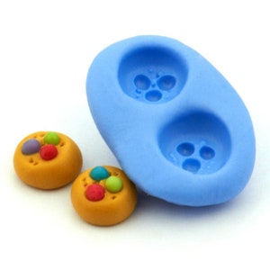 Könnte beinhalten: Eine blaue Silikonform mit zwei runden Vertiefungen zum Herstellen von Keksen. Zwei Kekse mit bunten Bonbons sind neben der Form zu sehen.