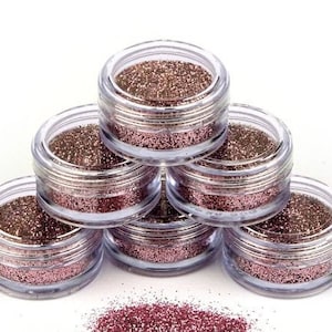 Op de afbeelding: Zes kleine, doorzichtige potjes gevuld met roze glitter, gestapeld in de vorm van een piramide. De potjes staan op een wit oppervlak.