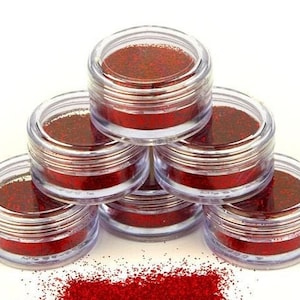 Op de afbeelding: Zes kleine, doorzichtige potjes gevuld met rode glitter, gestapeld in een piramidevorm. Een kleine hoop rode glitter is verspreid voor de potjes.