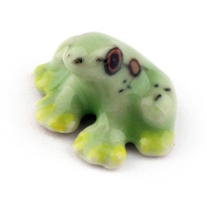 Può includere: Una piccola statuina di rana in ceramica verde con accenti gialli e occhi dipinti di marrone. La rana è seduta sulla pancia con le gambe distese.