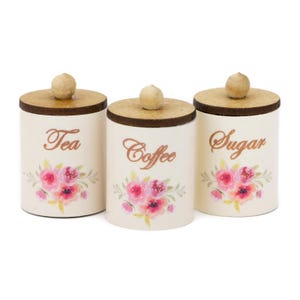 Könnte beinhalten: Drei weiße Keramikdosen mit Holzdeckeln und Blumendesigns. Die Dosen sind mit "Tee", "Kaffee" und "Zucker" beschriftet.