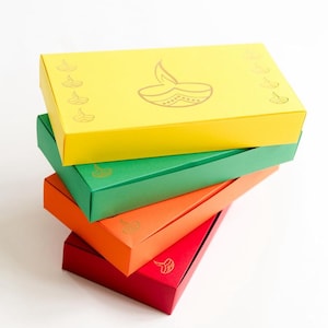 May include: A stack of colorful rectangular gift boxes in yellow, green, orange, and red. Each box features a gold-colored illustration of a lit diya lamp. The boxes are arranged in a diagonal stack, with the yellow box on top.