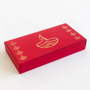 Könnte beinhalten: Eine rechteckige rote Geschenkbox mit einem goldfarbenen Diya-Design (Öllampe) auf dem Deckel. Die Box hat ein schlichtes, elegantes Design mit kleineren Diya-Motiven an den Rändern. Die Box ist wahrscheinlich zum Verschenken oder für besondere Anlässe gedacht.