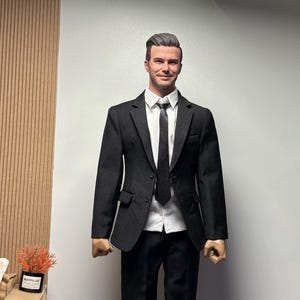 May include: A black suit, white shirt, and black tie. The figure wears black dress shoes. The background is neutral. The suit is a classic formal outfit.