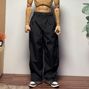 May include: A headless action figure wearing black pants with a drawstring waist and sneakers with brown and white accents. The figure has a muscular torso and arms. The background includes a plant, a couch, and a wooden floor.