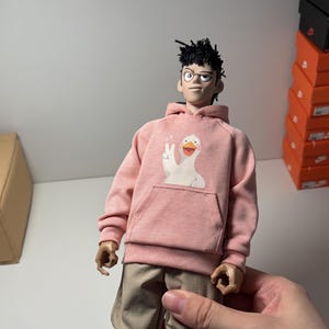 Peut inclure: Une poupée vêtue d'un sweat à capuche rose avec un motif d'oie blanche, d'un pantalon kaki et de baskets noires et blanches. La poupée a des cheveux noirs et des lunettes. La poupée est tenue par une main.