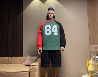 May include: A green, black, and red long-sleeved shirt with the number "84" printed on the front. The shirt is worn by a doll with black hair and a silver necklace. The doll is wearing black shorts and black and white sneakers.