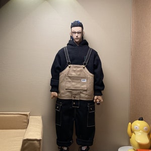 May include: A person wearing a black hoodie with a beige overall on top. The overall has a large pocket on the front and two smaller pockets on the sides. The person is wearing black pants and black and white sneakers.