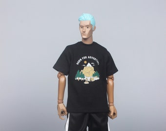 Puede incluir: Una camiseta negra con un gráfico de una cordillera y el texto "Born for Adventure". El modelo lleva pantalones negros con rayas blancas a los lados y zapatillas negras y verdes.