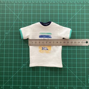 May include: White t-shirt with a blue and green striped graphic that says "LOVE" and a yellow rectangle with the text "1984". The shirt is measured with a ruler, showing it is 12 cm wide.