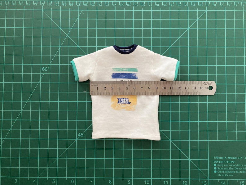 May include: White t-shirt with a blue and green striped graphic that says "LOVE" and a yellow rectangle with the text "1984". The shirt is measured with a ruler, showing it is 12 cm wide.
