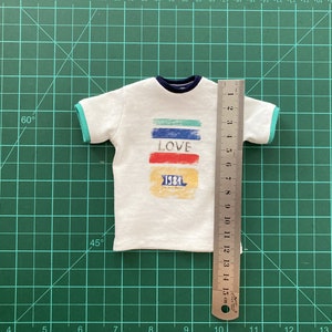May include: White t-shirt with a colorful graphic design that says "LOVE" and "1984". The shirt has a blue and green trim around the neck and sleeves. The shirt is measured with a ruler that shows it is 15 cm long.