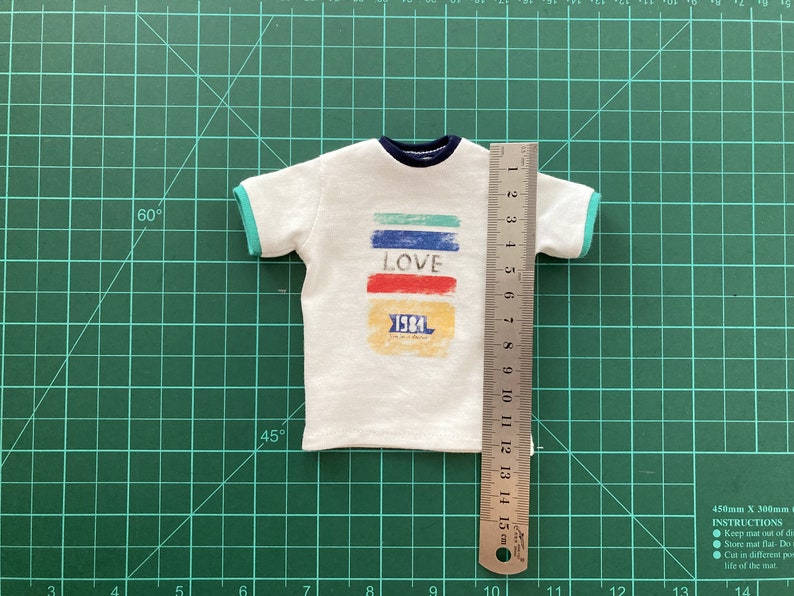 May include: White t-shirt with a colorful graphic design that says "LOVE" and "1984". The shirt has a blue and green trim around the neck and sleeves. The shirt is measured with a ruler that shows it is 15 cm long.