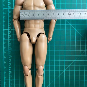 May include: A nude-colored plastic action figure with articulated limbs. The figure is standing on a green gridded mat with a ruler measuring 13 cm in length.