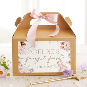 Puede incluir: Una caja de regalo de cartón marrón con un lazo de cinta rosa. La caja tiene una etiqueta que dice "Adeline's fairy first birthday" con ilustraciones de hadas y detalles florales. Una varita de estrella dorada está al frente.