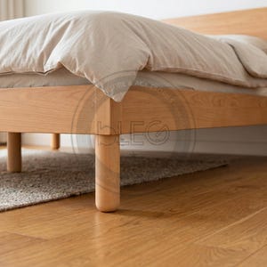 Könnte beinhalten: Ein hellfarbiger Holzbettenrahmen mit einem Kopfteil und vier abgerundeten Beinen. Das Bett ist mit einer beigen Bettdecke und Kissen bedeckt. Das Bett steht auf einem hellbraunen Hartholzboden, darunter ist ein grauer Teppich zu sehen.