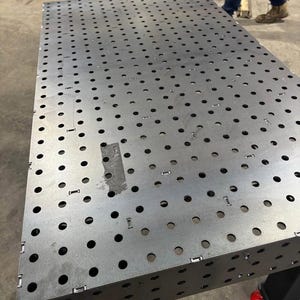 K&ouml;nnte beinhalten: Ein gro&szlig;er, rechteckiger Metall-Schwei&szlig;tisch mit zahlreichen, gleichm&auml;&szlig;ig verteilten L&ouml;chern. Der Tisch besteht aus silberfarbenem Metall und hat ein robustes, industrielles Design. Der Tisch wird von einem schwarzen Rahmen mit R&auml;dern getragen.