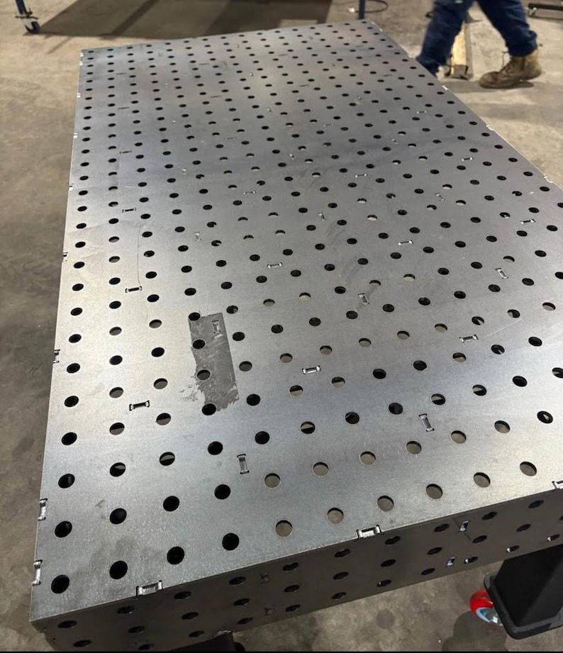 K&ouml;nnte beinhalten: Ein gro&szlig;er, rechteckiger Metall-Schwei&szlig;tisch mit zahlreichen, gleichm&auml;&szlig;ig verteilten L&ouml;chern. Der Tisch besteht aus silberfarbenem Metall und hat ein robustes, industrielles Design. Der Tisch wird von einem schwarzen Rahmen mit R&auml;dern getragen.