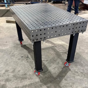 K&ouml;nnte beinhalten: Ein gro&szlig;er, rechteckiger Metall-Schwei&szlig;tisch mit zahlreichen, gleichm&auml;&szlig;ig verteilten L&ouml;chern. Der Tisch hat eine dunkelgraue Oberfl&auml;che und wird von vier schwarzen Beinen getragen. Jedes Bein ist mit einer rot-bereiften Rolle f&uuml;r Mobilit&auml;t ausgestattet. Der Tisch ist f&uuml;r den industriellen Einsatz konzipiert.