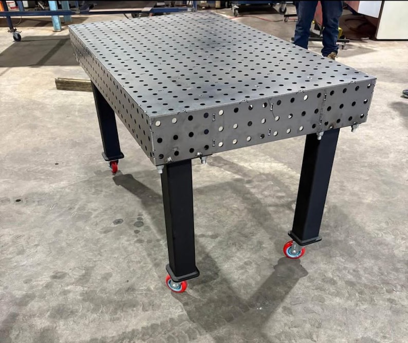 K&ouml;nnte beinhalten: Ein gro&szlig;er, rechteckiger Metall-Schwei&szlig;tisch mit zahlreichen, gleichm&auml;&szlig;ig verteilten L&ouml;chern. Der Tisch hat eine dunkelgraue Oberfl&auml;che und wird von vier schwarzen Beinen getragen. Jedes Bein ist mit einer rot-bereiften Rolle f&uuml;r Mobilit&auml;t ausgestattet. Der Tisch ist f&uuml;r den industriellen Einsatz konzipiert.