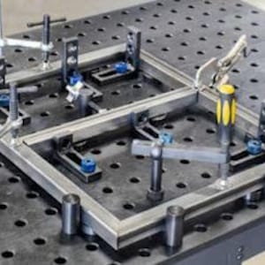 K&ouml;nnte beinhalten: Ein Metall-Schwei&szlig;tisch mit einem eingespannten Rahmen. Der Tisch hat eine dunkelgraue Oberfl&auml;che mit gleichm&auml;&szlig;ig verteilten L&ouml;chern. Klemmen und Halterungen halten den Metallrahmen, mit einem Werkzeug mit gelb-schwarzem Griff sichtbar.