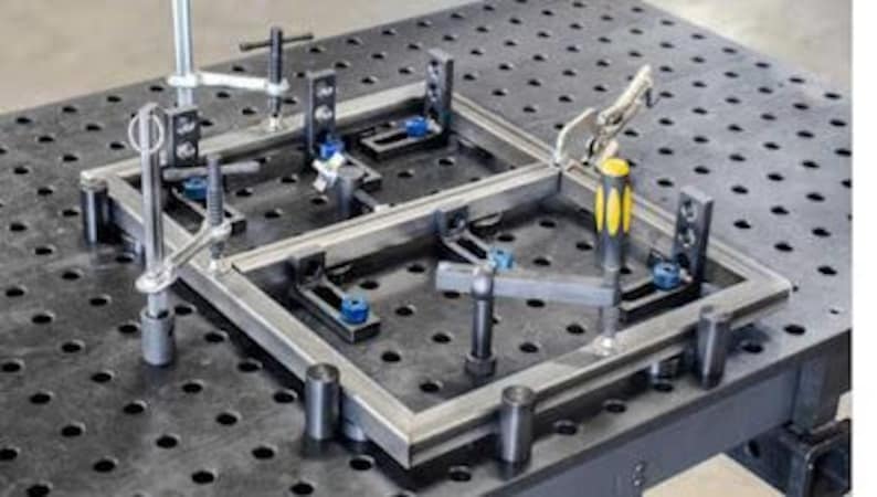 K&ouml;nnte beinhalten: Ein Metall-Schwei&szlig;tisch mit einem eingespannten Rahmen. Der Tisch hat eine dunkelgraue Oberfl&auml;che mit gleichm&auml;&szlig;ig verteilten L&ouml;chern. Klemmen und Halterungen halten den Metallrahmen, mit einem Werkzeug mit gelb-schwarzem Griff sichtbar.