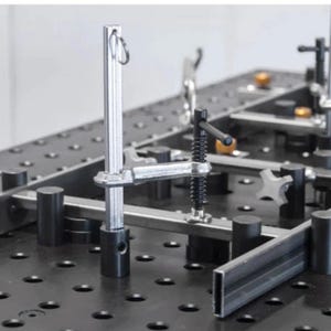 K&ouml;nnte beinhalten: Ein Metall-Schwei&szlig;tisch mit verschiedenen Klemmen und Vorrichtungen. Der Tisch hat eine schwarze Oberfl&auml;che mit zahlreichen L&ouml;chern, und Metallstangen werden durch silberne und schwarze Klemmen fixiert. Das Setup deutet auf eine Werkstattumgebung hin.