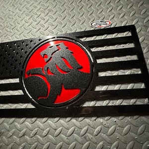 May include: Black metal wall art featuring a stylized American flag design. The flag incorporates a red circle with a lion emblem. The stars and stripes are cut out, revealing the background. The overall design is a patriotic and automotive-themed piece.