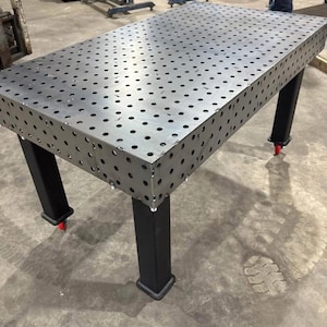 K&ouml;nnte beinhalten: Ein gro&szlig;er, rechteckiger Metall-Schwei&szlig;tisch mit zahlreichen, gleichm&auml;&szlig;ig verteilten L&ouml;chern auf der Oberfl&auml;che. Der Tisch hat schwarze Beine und kleine rote Rollen an der Unterseite. F&uuml;r den industriellen Einsatz konzipiert.