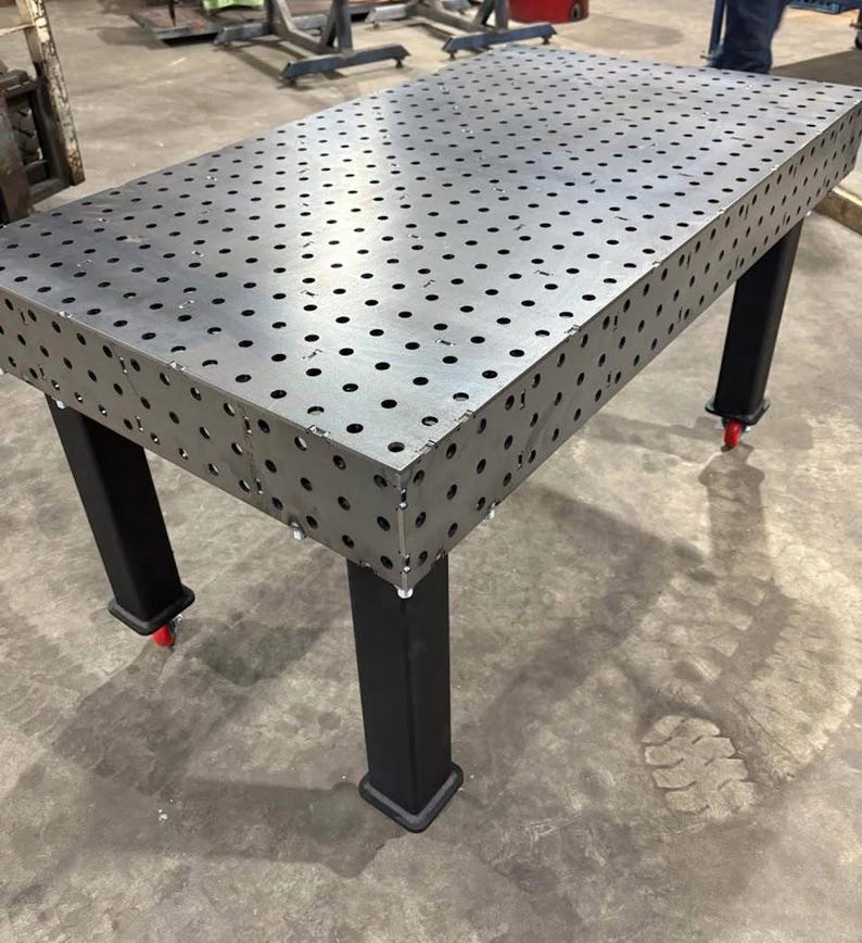 K&ouml;nnte beinhalten: Ein gro&szlig;er, rechteckiger Metall-Schwei&szlig;tisch mit zahlreichen, gleichm&auml;&szlig;ig verteilten L&ouml;chern auf der Oberfl&auml;che. Der Tisch hat schwarze Beine und kleine rote Rollen an der Unterseite. F&uuml;r den industriellen Einsatz konzipiert.